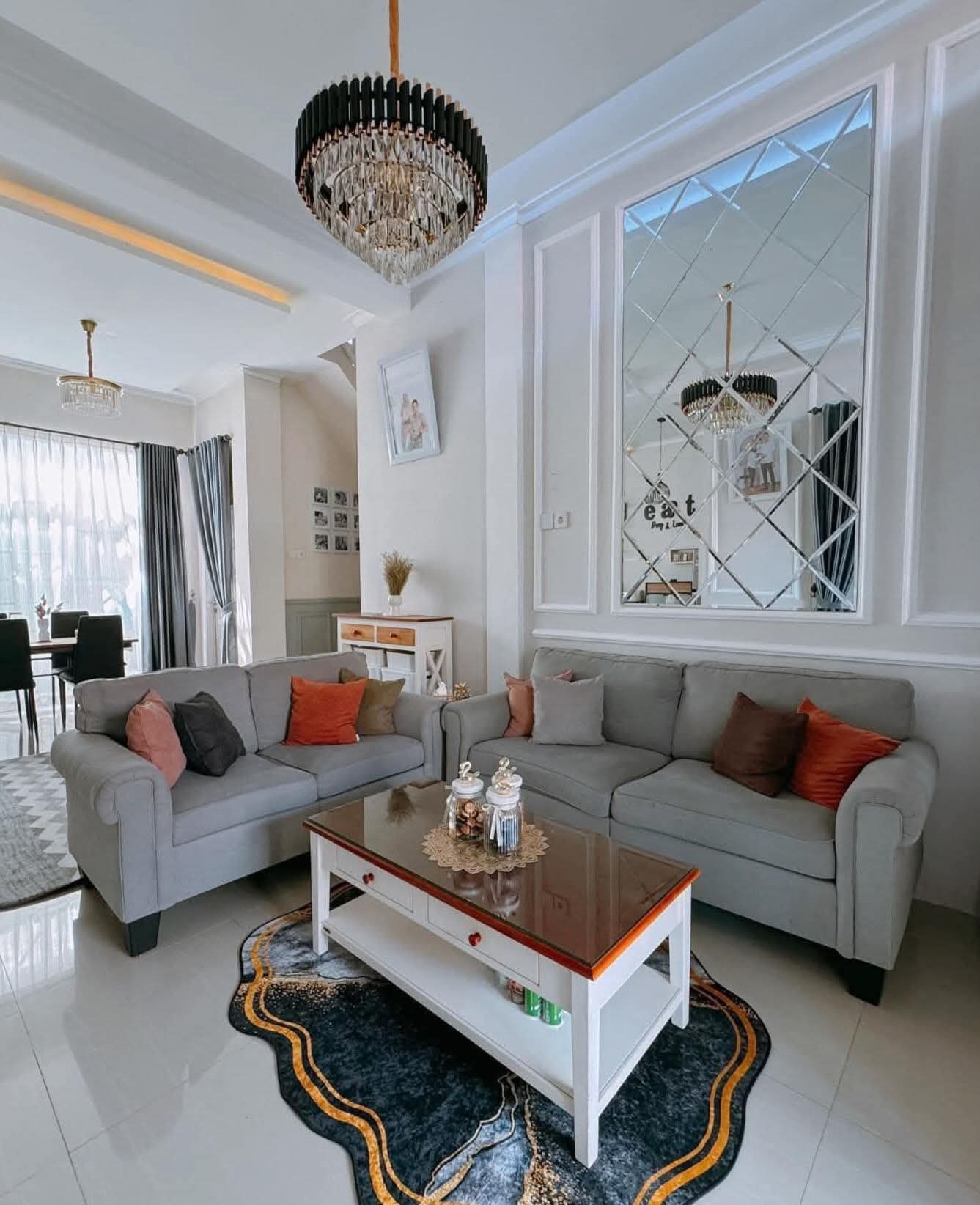 Ruang tamu modern dengan sofa abu-abu, bantal oranye, meja putih, dan lampu gantung kristal.