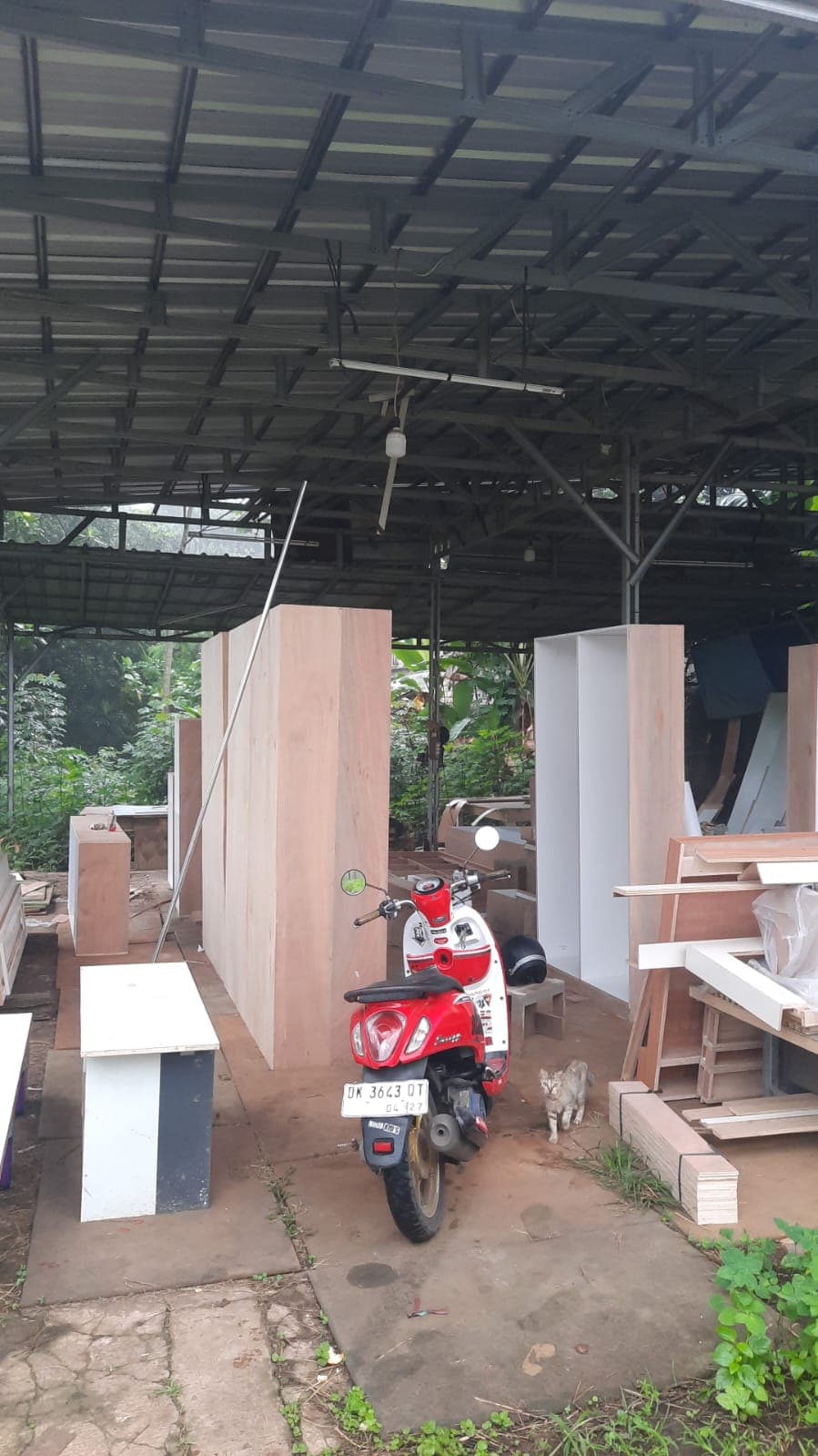 Sepeda motor merah terparkir di bengkel kayu dengan lemari besar dan seekor kucing kecil.