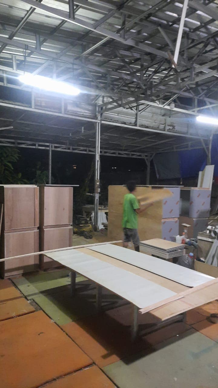 Pekerja berbaju hijau memindahkan papan kayu di dalam bengkel furnitur pada malam hari.